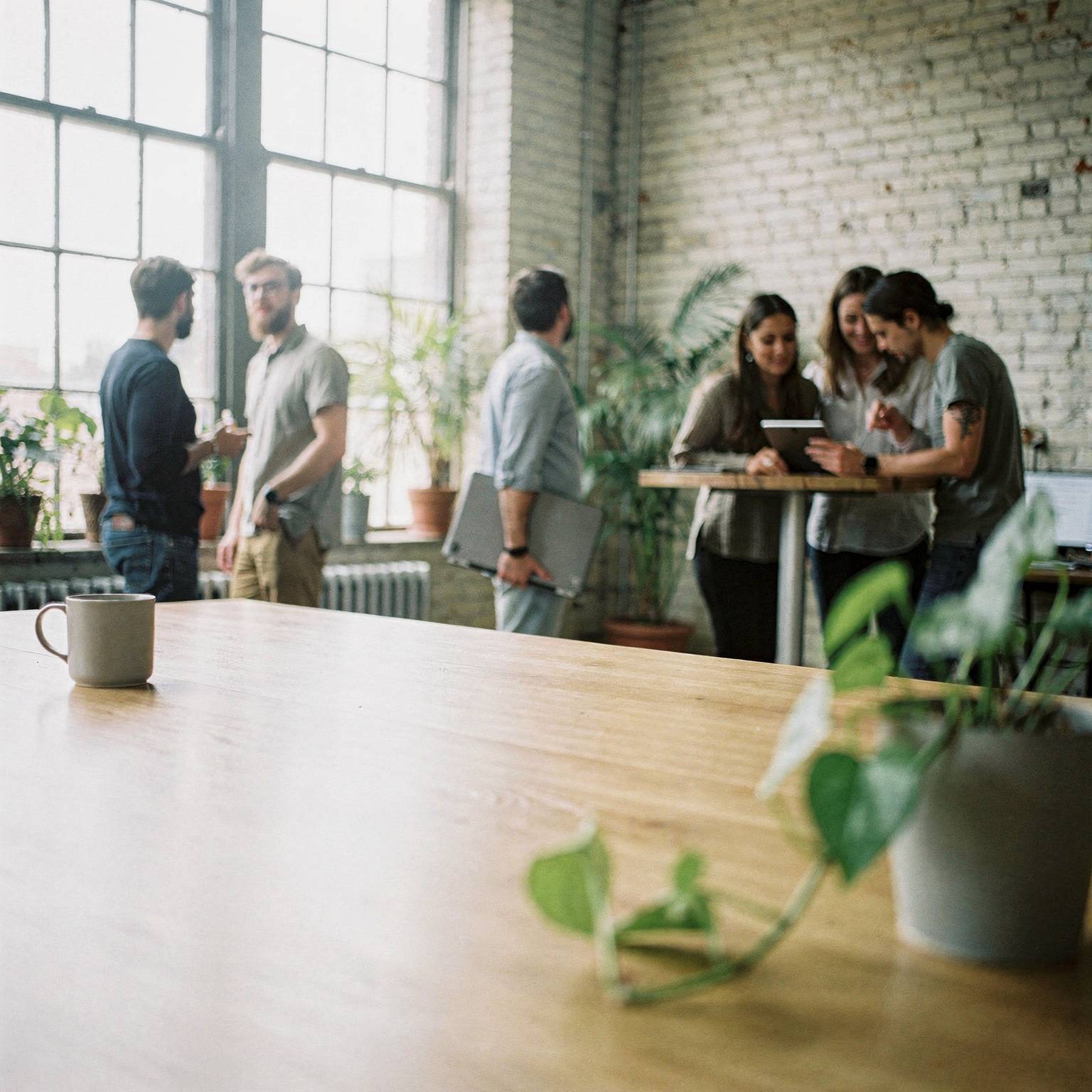 Colleagues in a bright loft office with brick walls, two talking by tall windows, one walking past with a laptop, and three gathered around a tablet at a standing table.