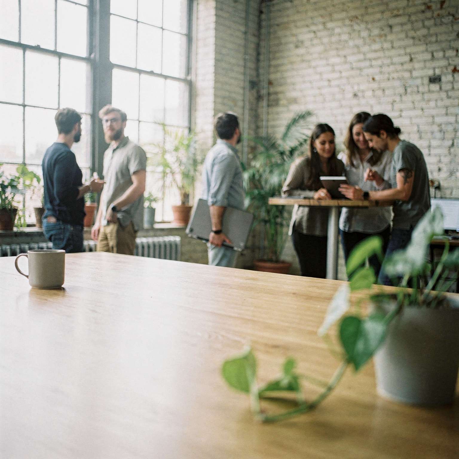 Colleagues in a bright loft office with brick walls, two talking by tall windows, one walking past with a laptop, and three gathered around a tablet at a standing table.