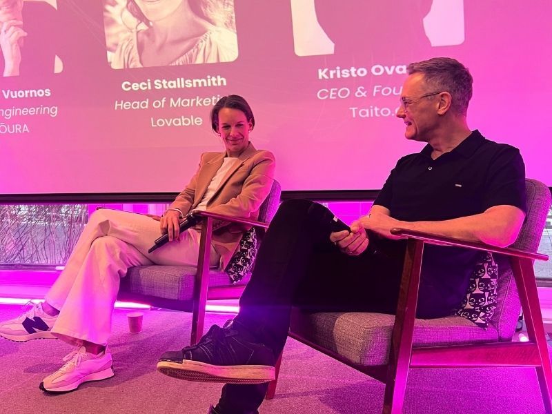 Two panelists seated on stage during a discussion on performance in the age of AI, with a presentation slide visible behind them and pink stage lighting at a Slush side event setting.